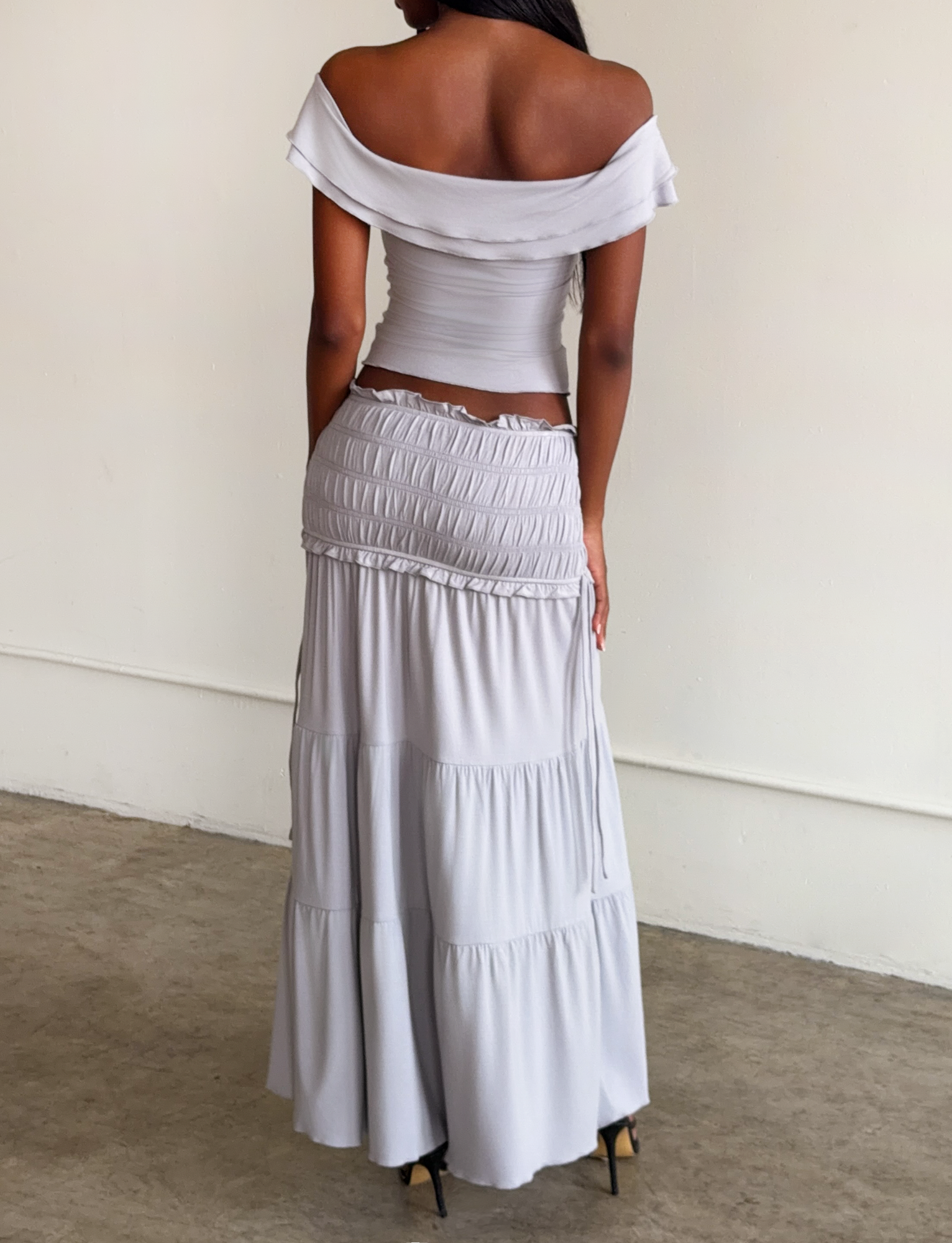 A woman stands indoors facing away, wearing the TIGERMIST DENALI MAXI SKIRT in Dove Grey with a shirred waistband and gathers, paired with an off-shoulder light gray fitted top and black high heels.