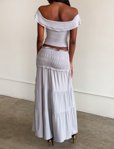 A woman stands indoors facing away, wearing the TIGERMIST DENALI MAXI SKIRT in Dove Grey with a shirred waistband and gathers, paired with an off-shoulder light gray fitted top and black high heels.