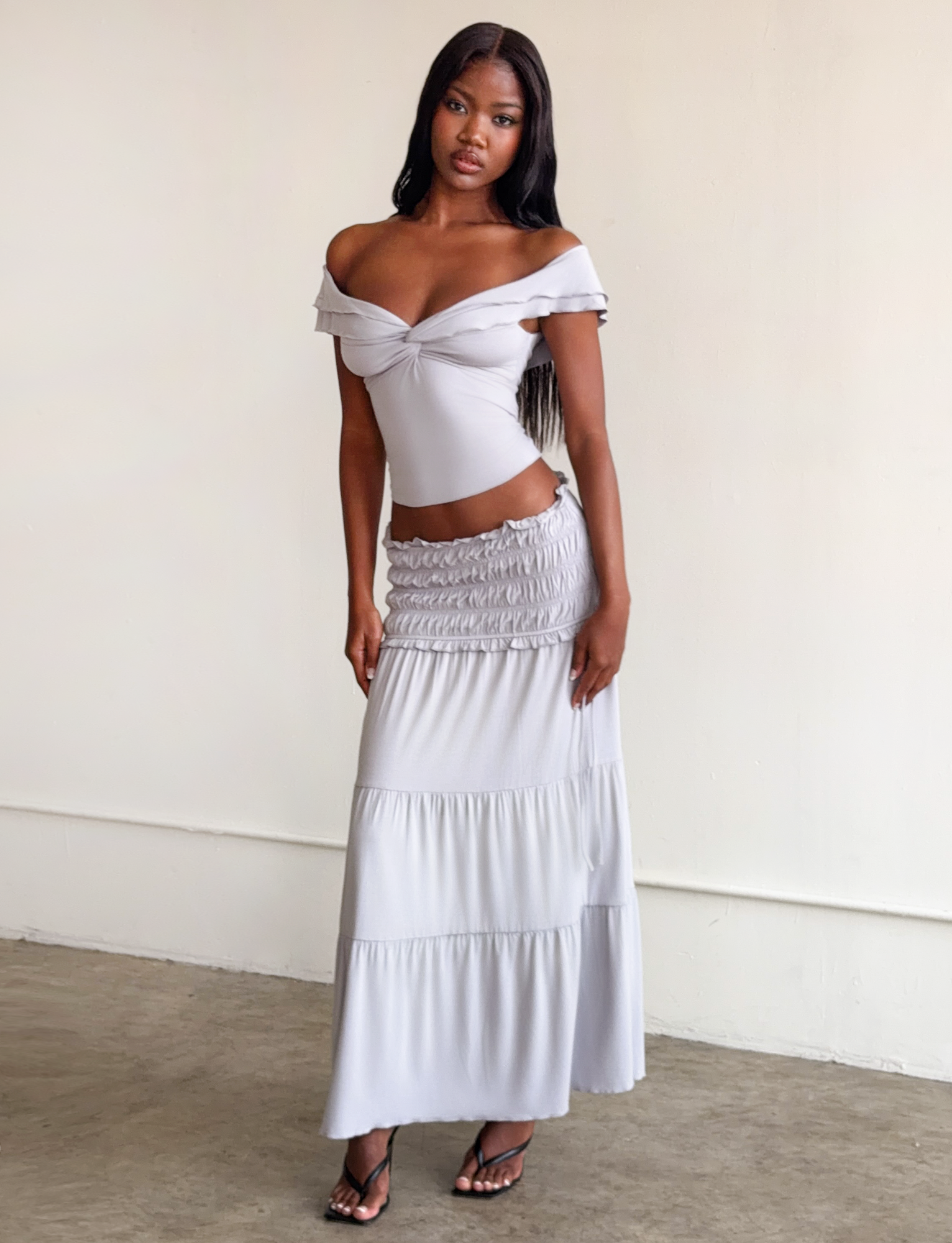 A woman stands indoors by a plain light wall, wearing the TIGERMIST DENALI TOP in dove grey with ruffled sleeves and a matching long tiered skirt. She has long dark hair and black heeled sandals.