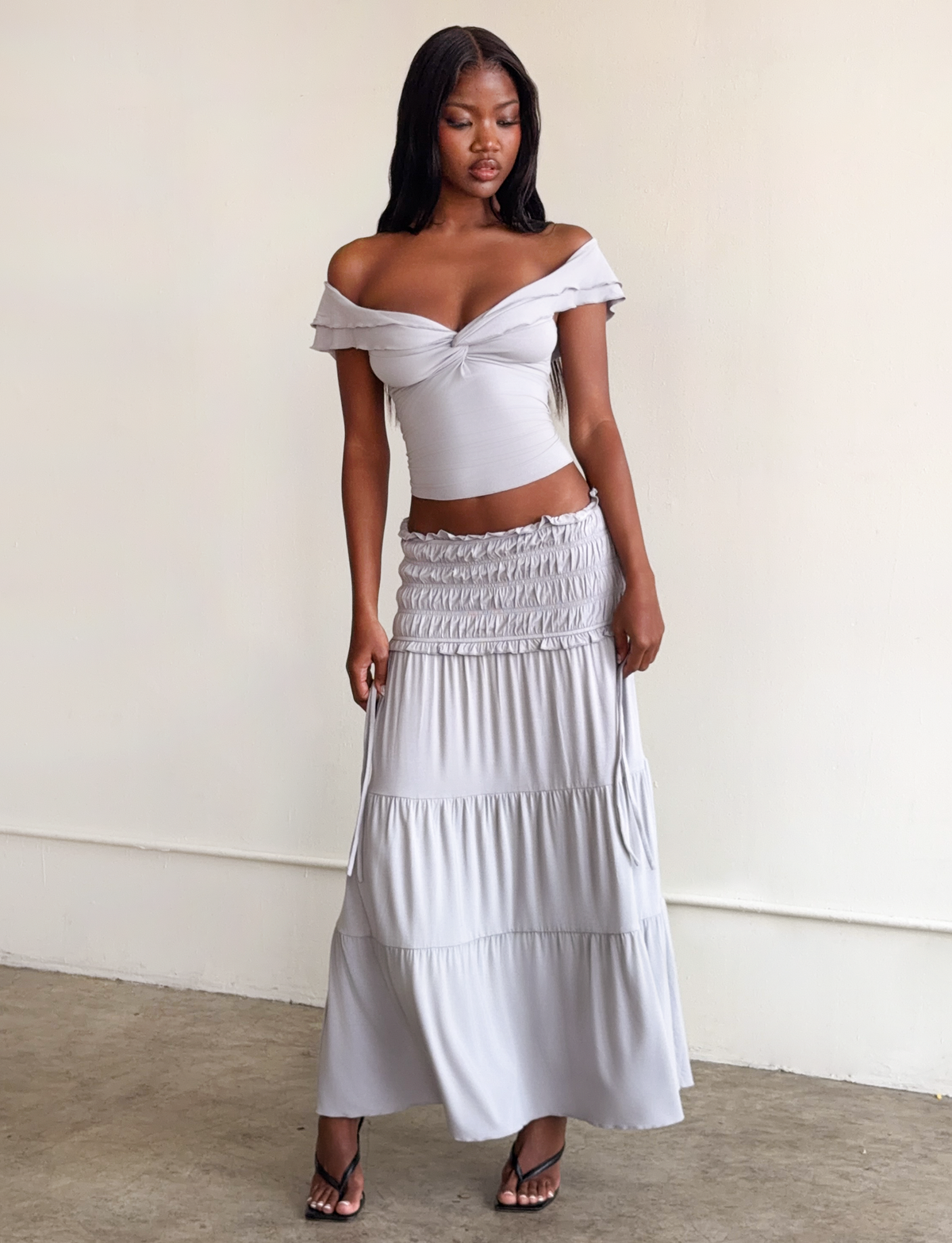 A woman stands indoors wearing the TIGERMIST DENALI TOP in dove grey, an off-the-shoulder style paired with a matching tiered maxi skirt made from soft modal fabric. She poses gracefully, one hand by her side and the other resting on her thigh.