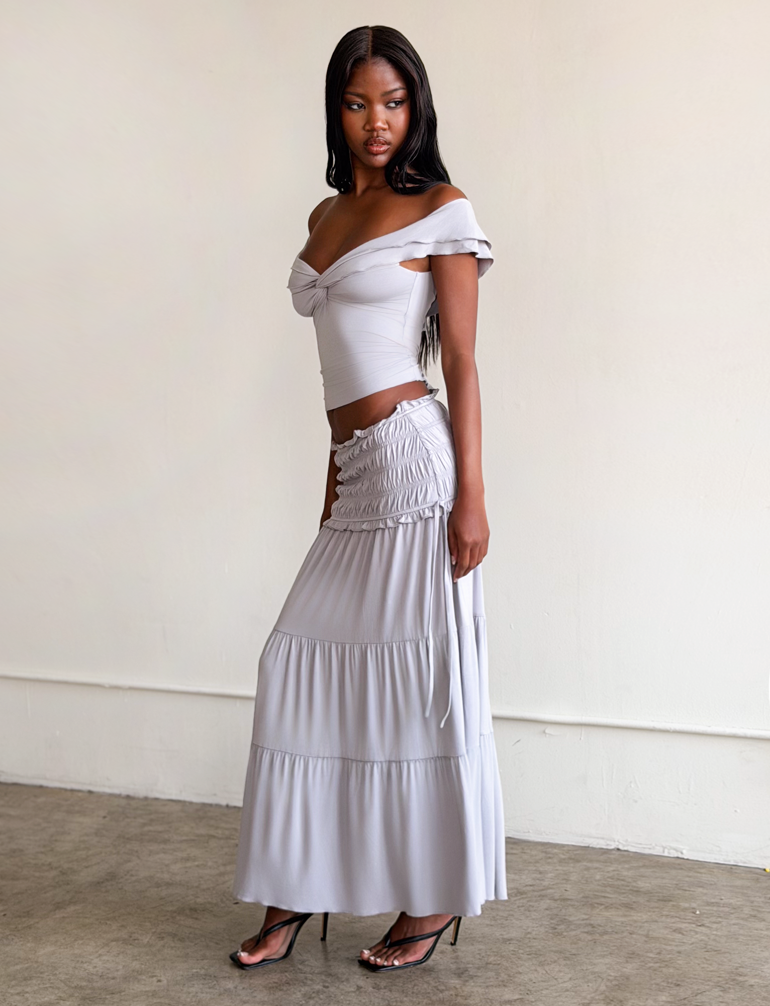 A woman stands indoors in a dove grey DENALI MAXI SKIRT by TIGERMIST, paired with a matching off-the-shoulder top. The soft modal skirt features a shirred waistband. She wears black heels and gazes to the side against a minimal background.
