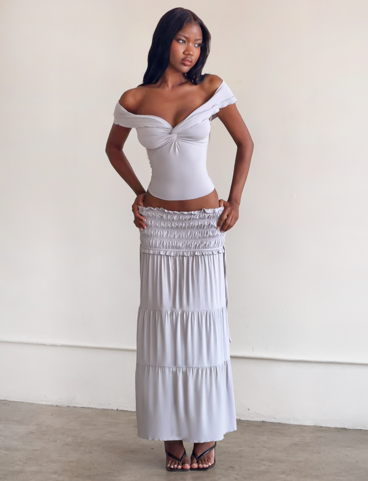 A woman stands indoors against a plain wall, wearing the TIGERMIST DENALI MAXI SKIRT in Dove Grey, featuring ruched details and a shirred waistband, paired with a light off-shoulder top and high-heeled sandals. She has long, straight hair.