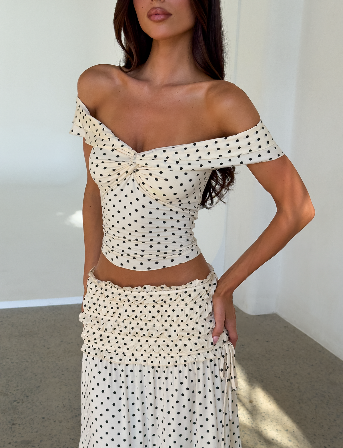 A woman wears the TIGERMIST DENALI TOP - CREAM DOT—an off-shoulder, fitted cream top with black polka dots—paired with a matching high-waisted ruffled skirt, standing indoors against a plain white wall.
