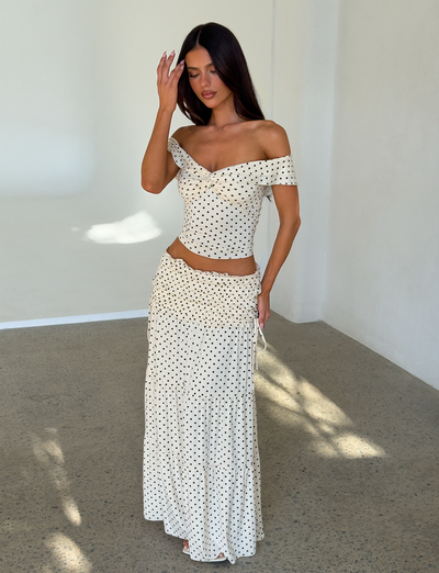 A woman stands indoors in a white off-the-shoulder crop top and the TIGERMIST DENALI MAXI SKIRT - CREAM DOT, both featuring black polka dots. She raises a hand to her head as sunlight fills the minimal, white-walled room.