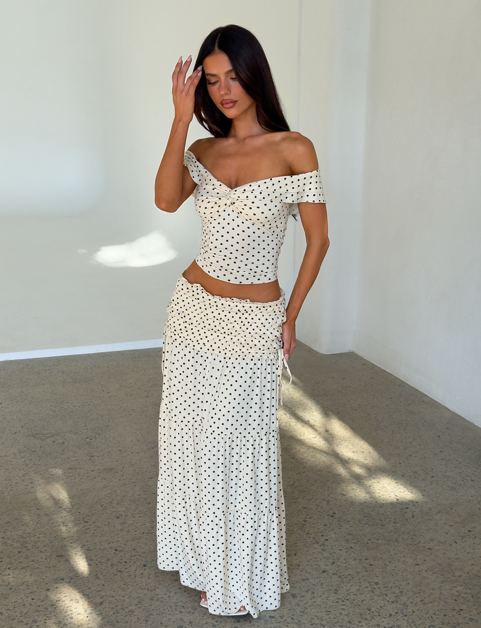 A woman stands indoors in a white off-the-shoulder crop top and the TIGERMIST DENALI MAXI SKIRT - CREAM DOT, both featuring black polka dots. She raises a hand to her head as sunlight fills the minimal, white-walled room.