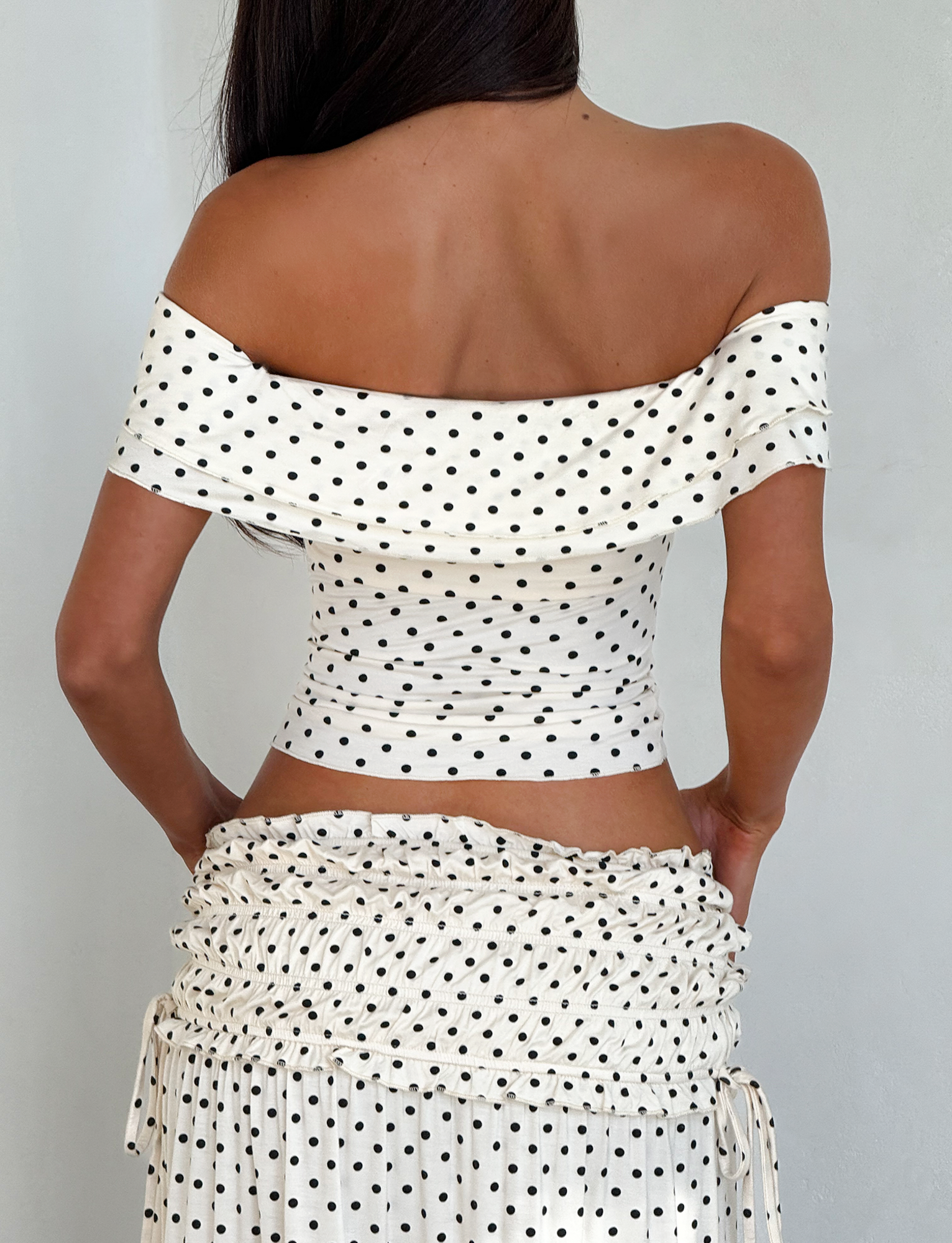 A woman with long dark hair wears a TIGERMIST DENALI MAXI SKIRT - CREAM DOT and a matching off-the-shoulder white top, both in soft modal fabric. Shown from the back, her hands are on her hips.