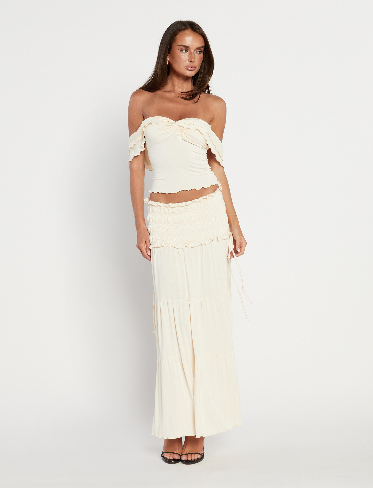 A woman stands in heeled sandals against a white background, wearing the TIGERMIST DENALI MAXI SKIRT in Bone with ruffle details and a flared silhouette, paired with a cream off-the-shoulder fitted top.