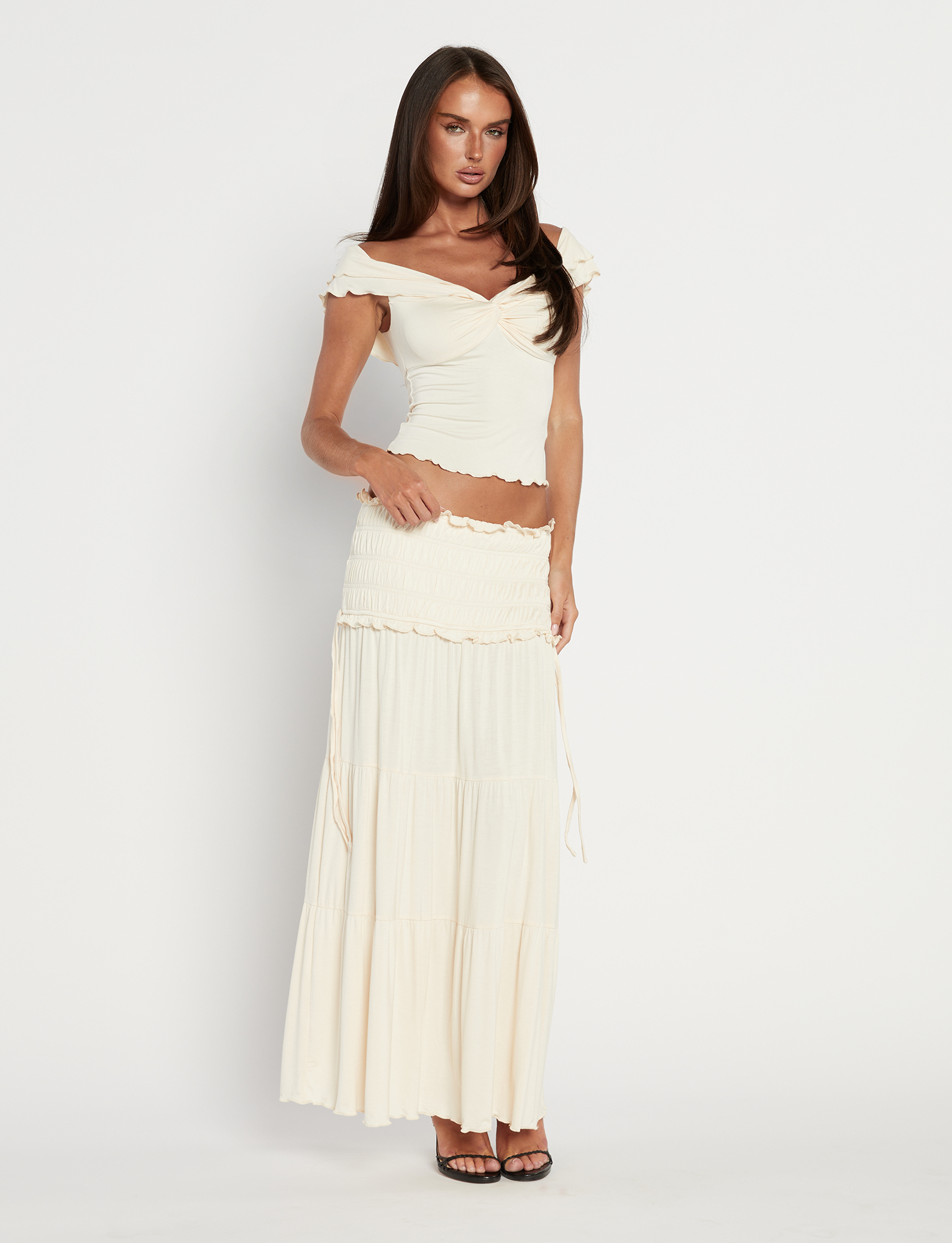 A woman with long brown hair poses against a white background wearing the TIGERMIST DENALI MAXI SKIRT - NEUTRAL : BONE, styled with an off-shoulder cream crop top and black high-heeled sandals. The skirt features a flared silhouette and ruffled edges.