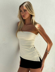 A woman with long blonde hair wears the TIGERMIST INDIRA TOP in cream, a strapless fitted piece, paired with black shorts. She stands against a white wall, gazing sideways with a serious look, her bent arm accessorized by two silver bangles.