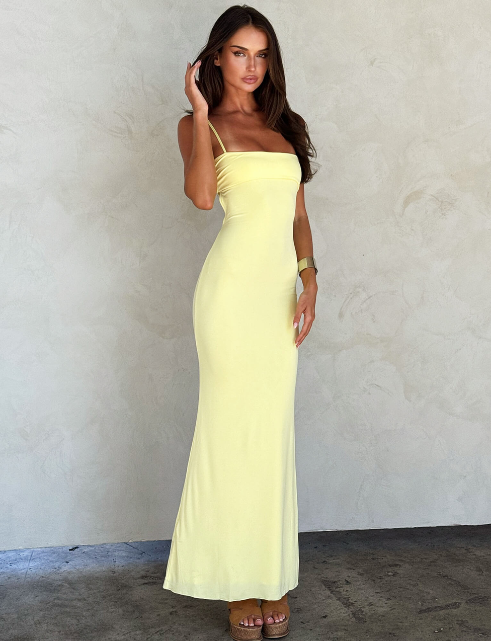 A woman poses gracefully against a light textured wall in the TIGERMIST CARA DRESS - YELLOW : PASTEL CITRUS, a pastel yellow bodycon bandeau dress, paired with brown platform sandals. She has long, wavy brown hair and one hand near her face.