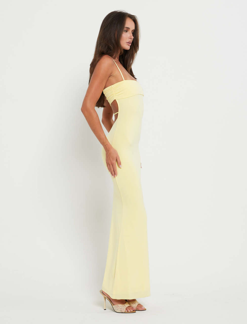A woman stands in profile against a white background wearing the TIGERMIST CARA DRESS - YELLOW : PASTEL CITRUS, a floor-length, sleeveless bodycon dress with thin straps, open sides, and paired with high-heeled sandals for an elegant look.