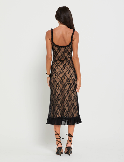 A woman with long dark hair, facing away, wears the TIGERMIST DEJA DRESS - BLACK: LACE, a sleeveless lace midi dress with a sheer pattern and ruffled hem. She pairs it with black strappy heels against a plain white background.