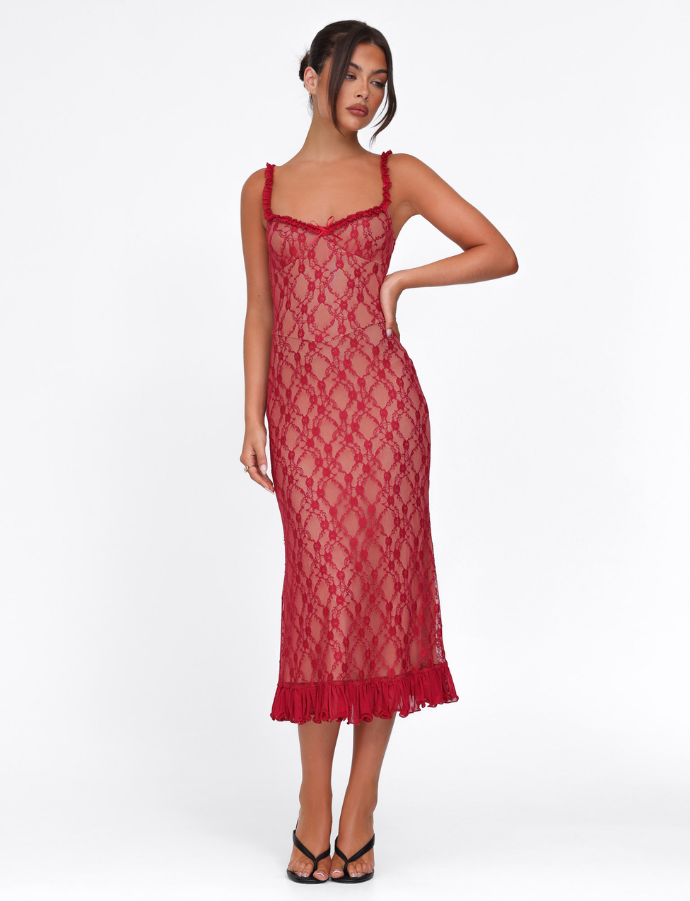 A woman poses against a white background wearing the TIGERMIST DEJA DRESS in Berry Red, a sleeveless, midi-length sheer lace dress with a ruffled hem, styled with black heeled sandals and her hair up, one hand on her hip.