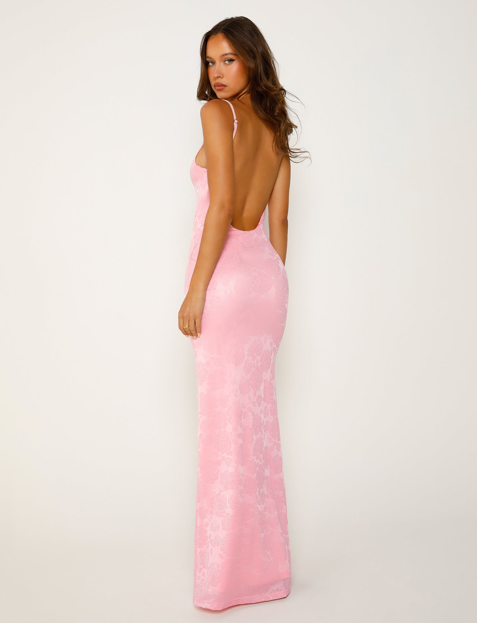 A person wearing the ERICKA DRESS - PINK from TIGERMIST poses against a plain, light-colored background. The long, form-fitting dress features a low back and a subtle floral pattern. The individual has long hair and is looking over their shoulder toward the camera.
