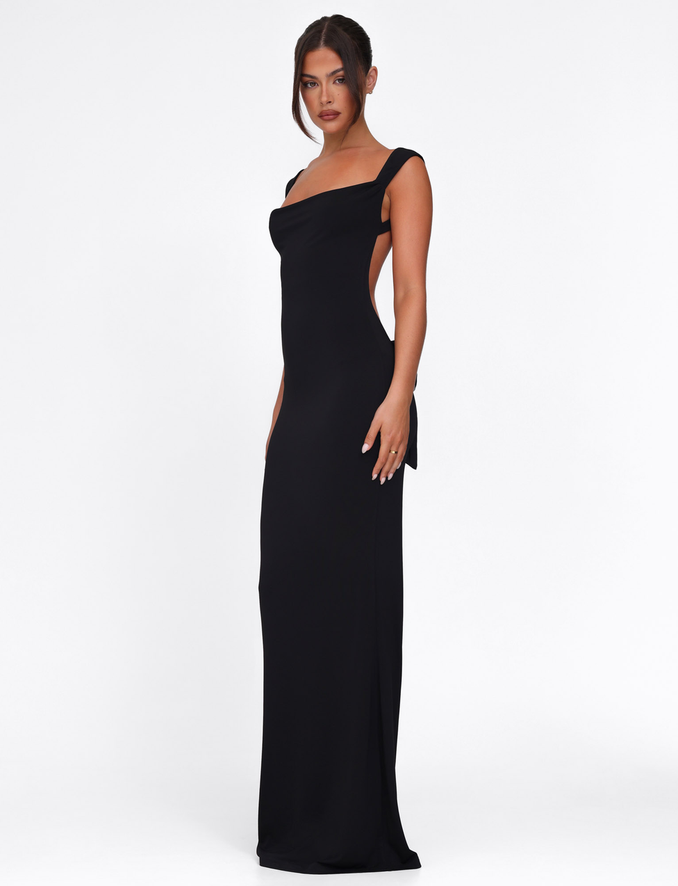 A woman poses confidently against a white background in the TIGERMIST ZITA DRESS - BLACK, featuring a bodycon silhouette, open back, and wide shoulder straps. Her hair is styled in a loose updo.