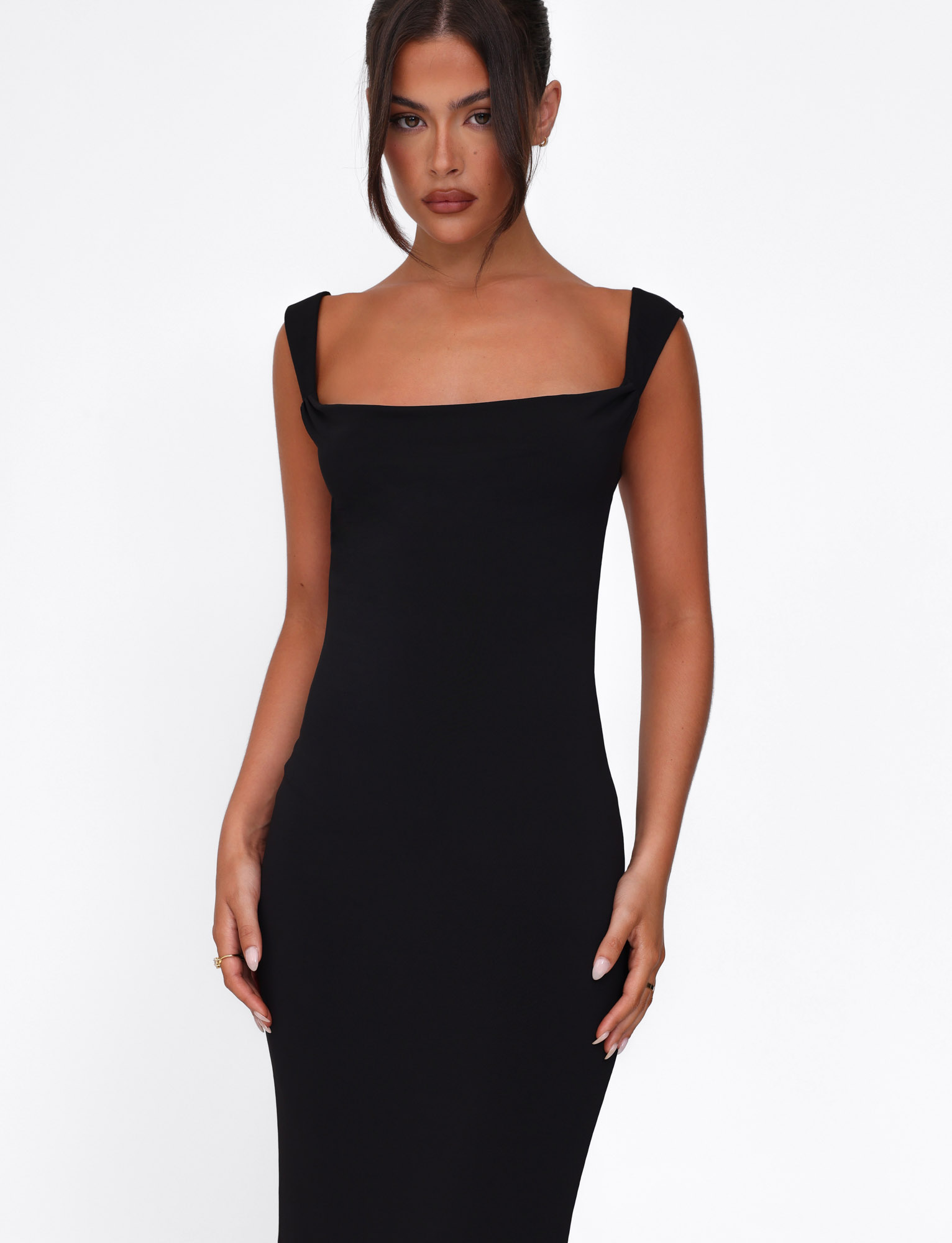 A woman with dark hair styled up wears the ZITA DRESS - BLACK by TIGERMIST, a fitted bodycon maxi dress with wide shoulder straps, standing against a plain white background.