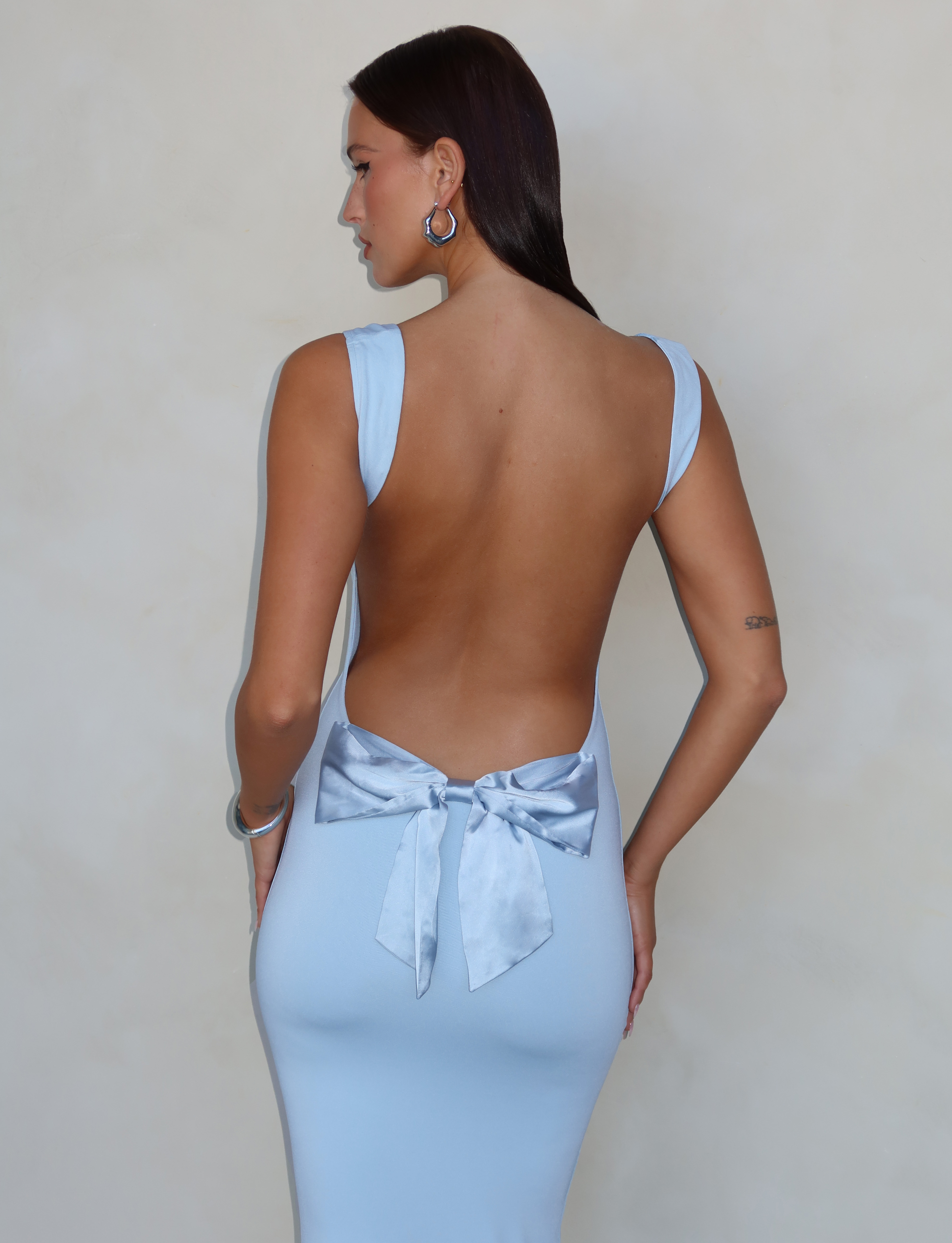 A woman with straight dark hair wears the TIGERMIST ZITA DRESS - BLUE: BABY BLUE, a backless bodycon style featuring a large satin bow at the lower back, as she stands facing away against a plain, neutral background.