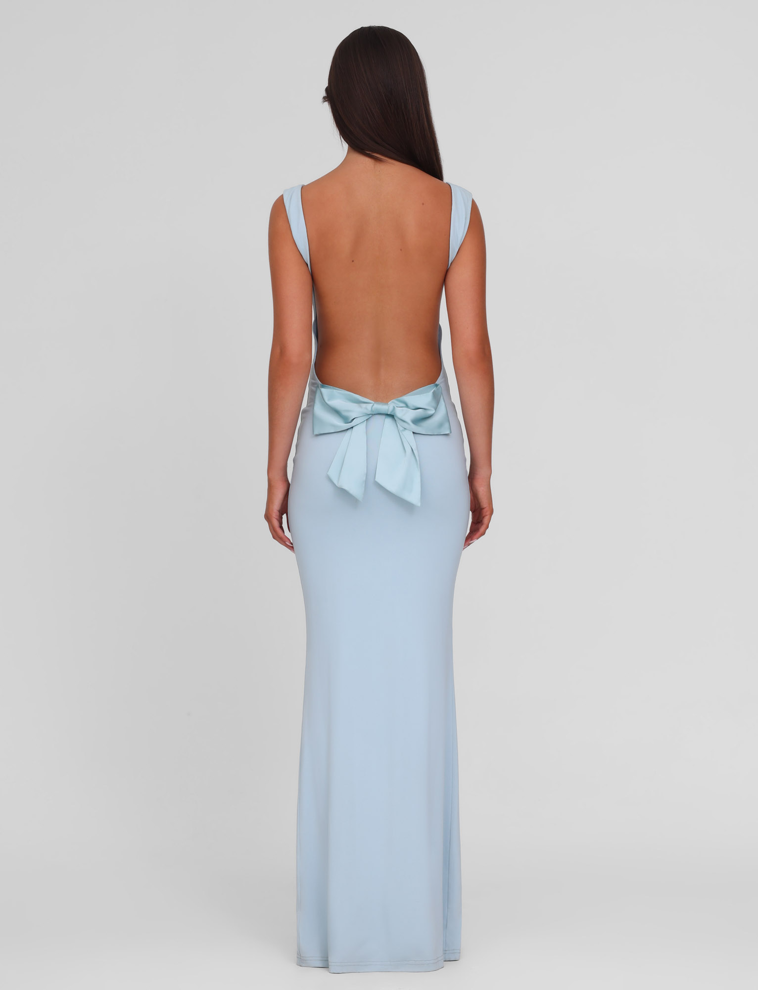 A woman with long brown hair models the TIGERMIST ZITA DRESS - BLUE : BABY BLUE, a light blue maxi-length backless gown with wide straps and a large bow at the lower back, standing against a plain white background.