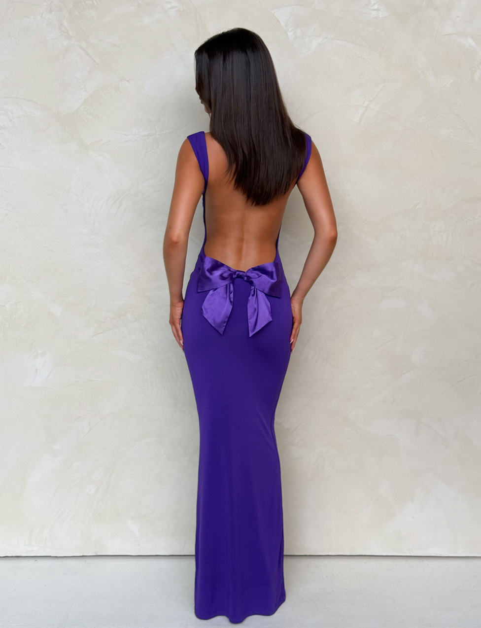 A woman with straight, dark hair wears the TIGERMIST ZITA MAXI DRESS in purple, featuring a bodycon silhouette and a statement bow at the lower back, standing against a light textured wall.