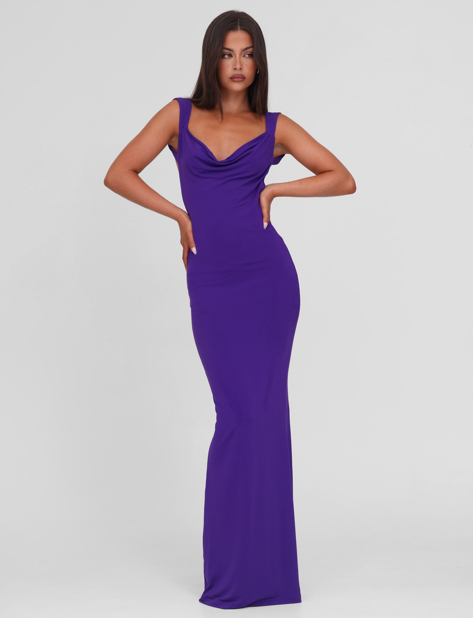 A woman poses confidently in the TIGERMIST ZITA MAXI DRESS - PURPLE, a sleeveless, bodycon, floor-length dress with a scoop cowl neck, standing against a plain white background.