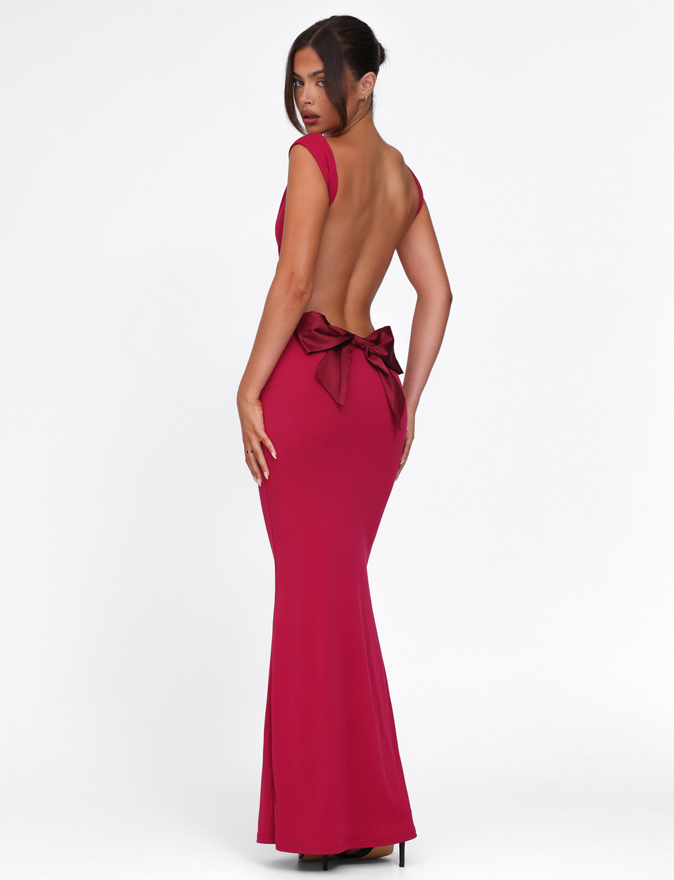 A woman in the TIGERMIST ZITA DRESS - RED, a floor-length, backless deep red bodycon gown with a large bow at the lower back, stands facing away and glances over her shoulder against a plain white background.