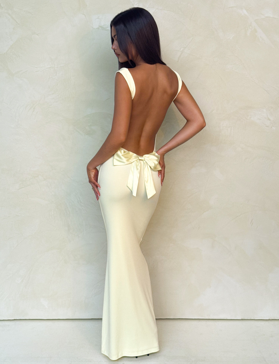 A woman wearing the Tiger Mist USA CL - ZITA MAXI DRESS, a long pale yellow gown with a backless design and large bow at the lower back, stands against a light textured wall, gazing to the side.