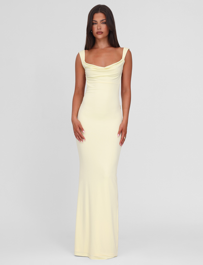 A woman with long dark hair wears the TIGERMIST ZITA MAXI DRESS in Lemon, featuring a scoop cowl neck and draped neckline, standing against a plain light gray background.