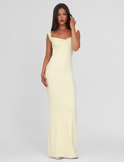A woman with long dark hair stands against a plain background wearing the TIGERMIST ZITA MAXI DRESS - LEMON, a sleeveless light yellow bodycon gown with a scoop cowl neck. She touches her face and gazes slightly to the side.