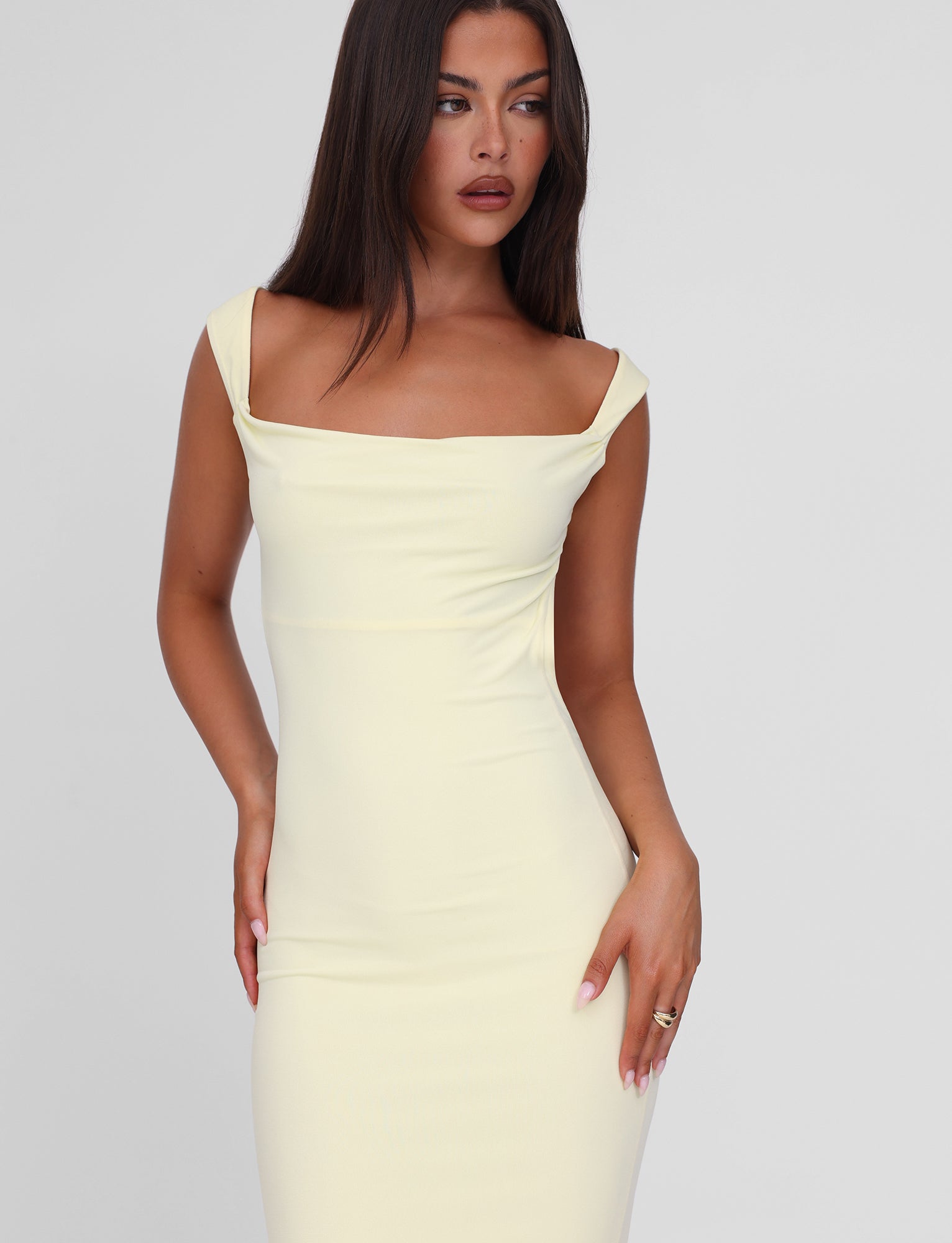 A woman with long, straight dark hair models the TIGERMIST ZITA MAXI DRESS in lemon. The fitted, off-shoulder bodycon dress stands out against a plain light gray backdrop as she poses looking to the side with one hand on her hip.