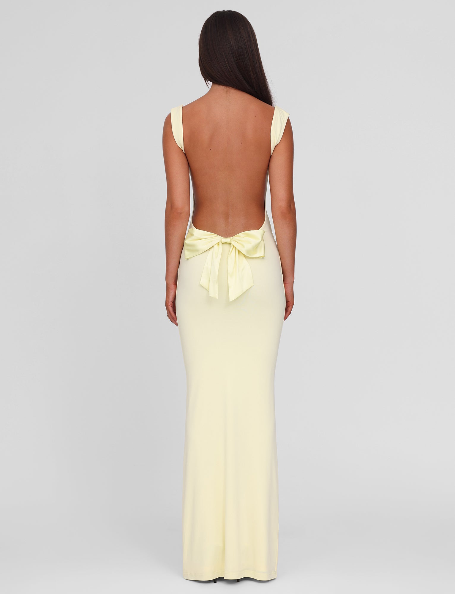 A woman with long brown hair faces away, wearing the TIGERMIST ZITA MAXI DRESS - LEMON—a pale yellow, backless, floor-length gown with wide straps, a large lower-back bow, and an elegant bodycon fit against a plain light gray background.
