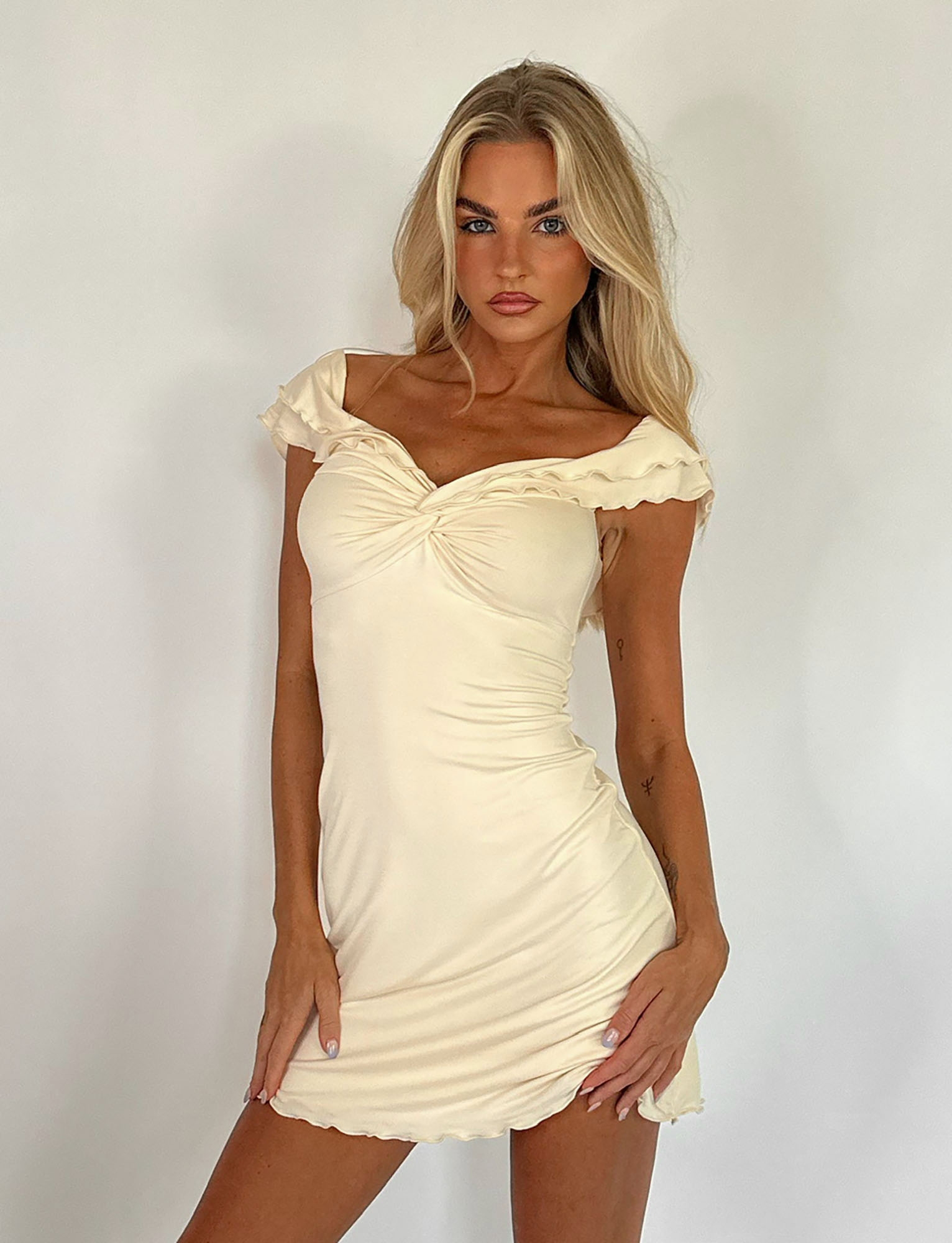 A woman with long blonde hair poses in the TIGERMIST DENALI MINI DRESS - WHITE : BONE, an off-the-shoulder, cream-colored mini dress, against a plain white background.