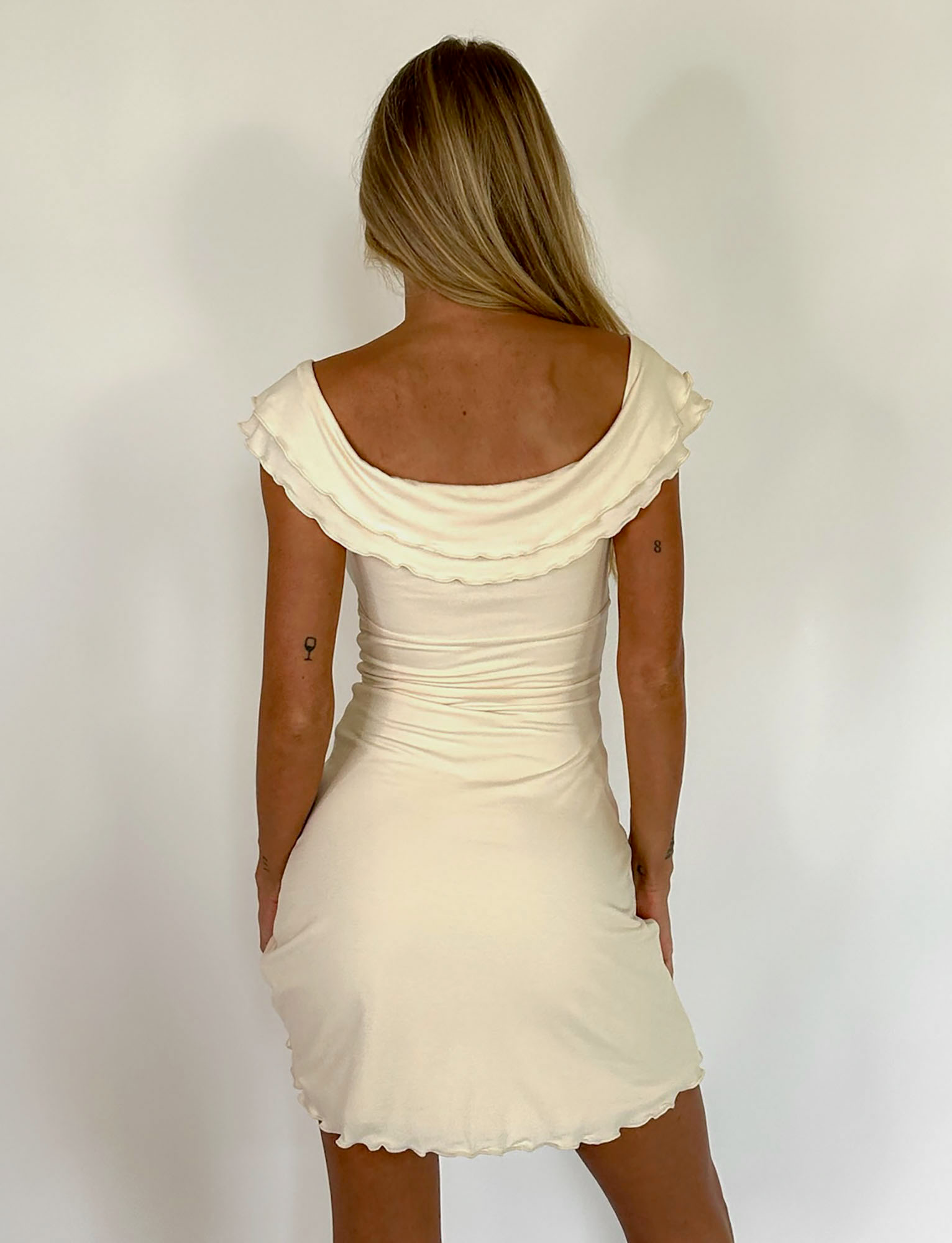 A woman with long blonde hair faces away, modeling the TIGERMIST DENALI MINI DRESS in White: Bone—an off-the-shoulder, cream mini dress with ruffled edges and a soft A-line shape—against a plain white background.
