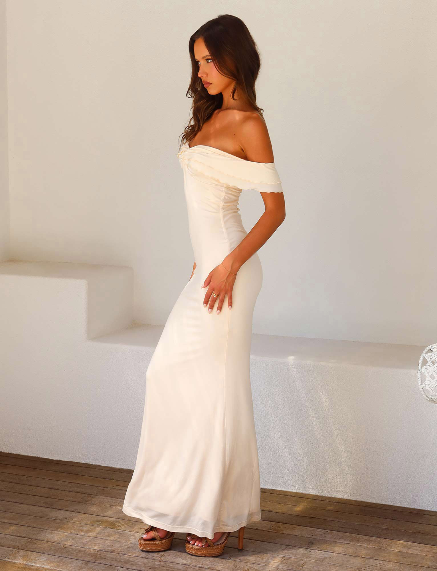 A woman with long brown hair wears the TIGERMIST DENALI MAXI DRESS - WHITE : BONE with a sweetheart neckline and platform sandals, standing indoors on a wooden floor against a plain white wall.