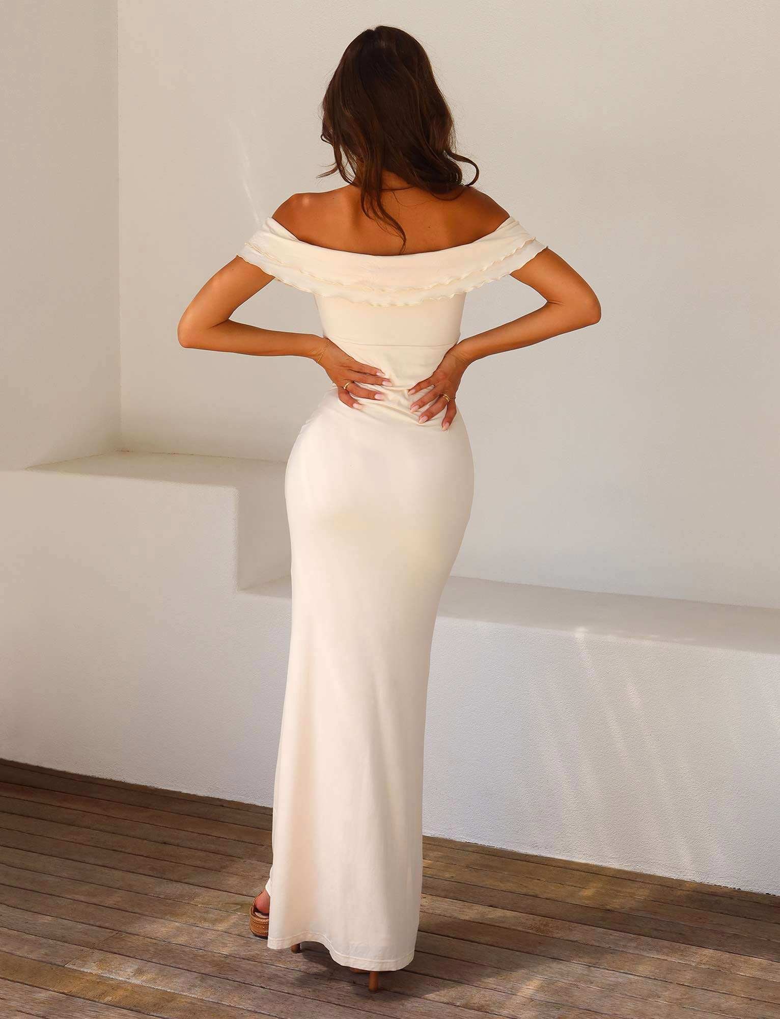 A woman with wavy brown hair stands indoors facing away, wearing the TIGERMIST DENALI MAXI DRESS - WHITE : BONE, an off-the-shoulder, sweetheart neckline bodycon maxi, paired with heeled sandals and hands resting on her lower back.