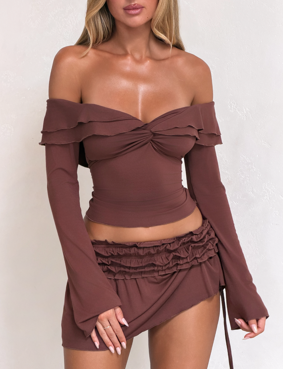 Woman in the Tiger Mist USA CL - DENALI LONG SLEEVE TOP, an off-shoulder, ruffled brown crop top with matching mini skirt, stands against a light textured background. Her face is partially out of frame.