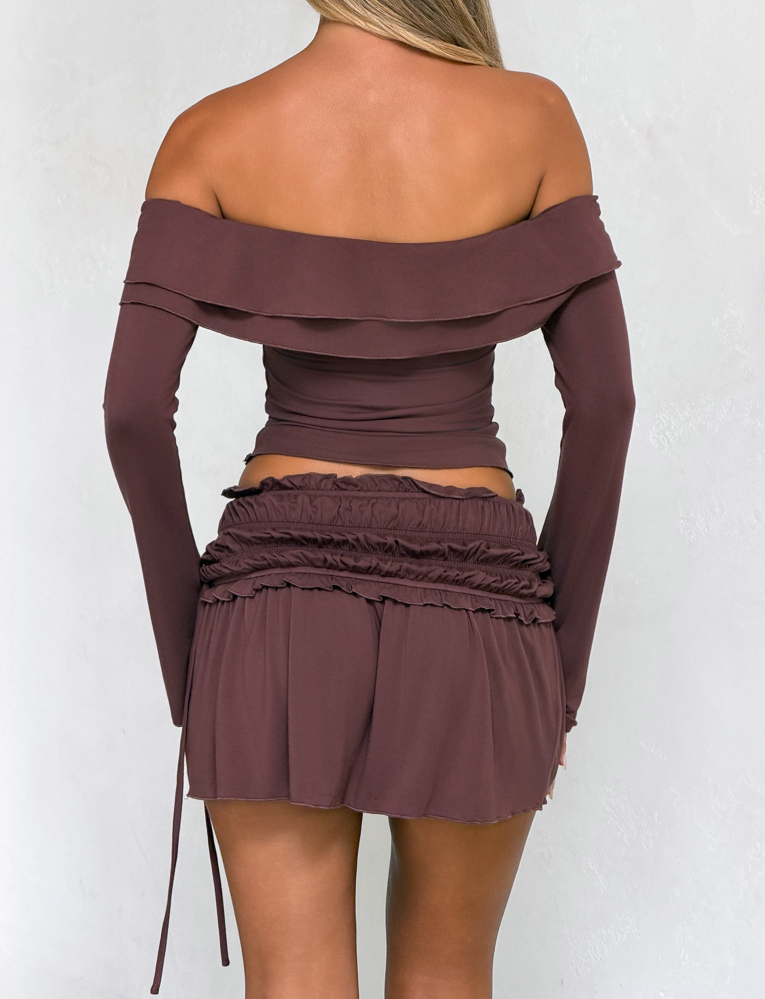 A woman stands with her back to the camera, wearing an off-shoulder top and the TIGERMIST DENALI MINI SKIRT - CHOCOLATE, which features ruffle details and a wide shirred waistband, against a light background.