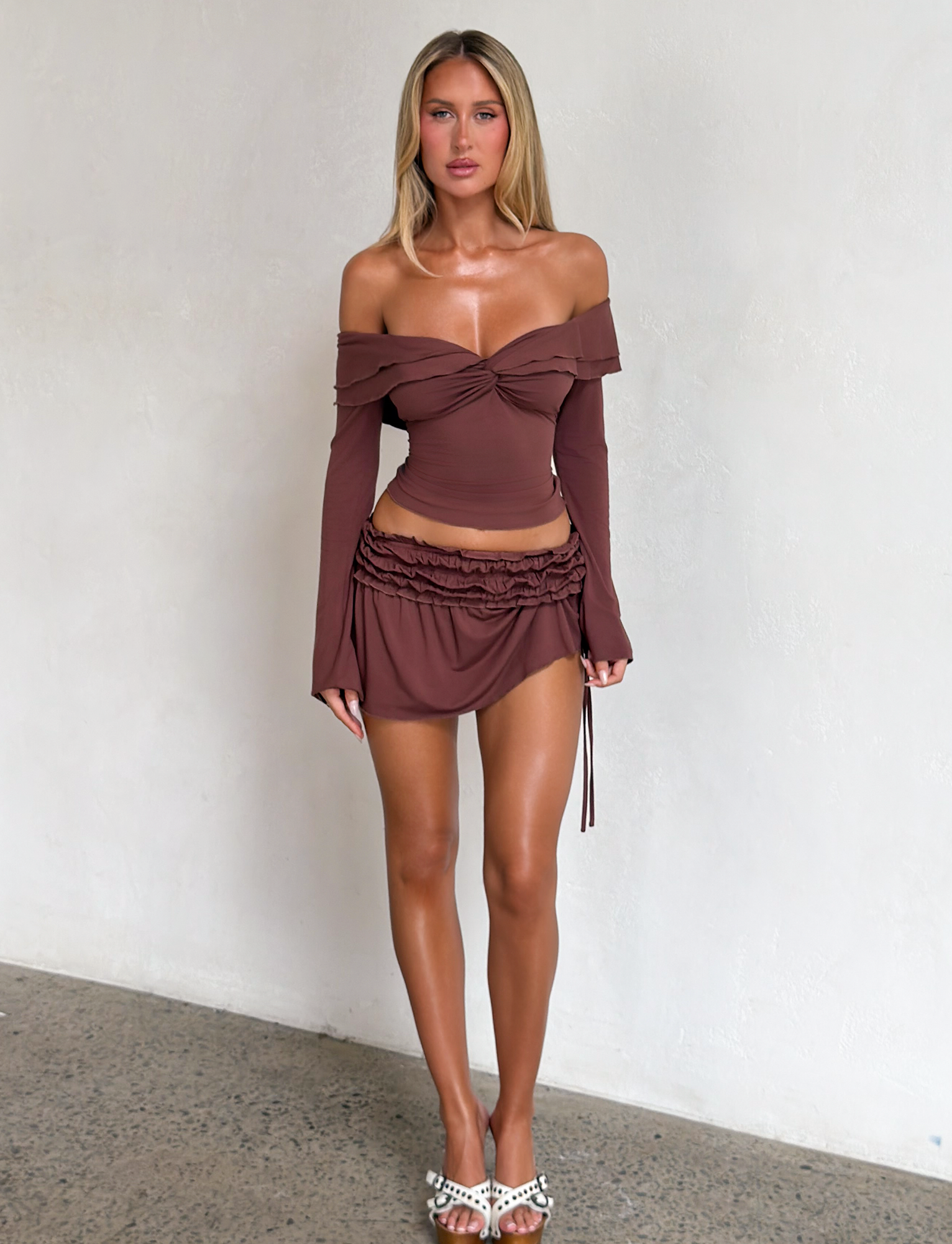 A woman with long blonde hair wears the TIGERMIST DENALI LONG SLEEVE TOP in chocolate with a matching mini skirt and white pearl-detailed heeled sandals, standing against a plain white wall.