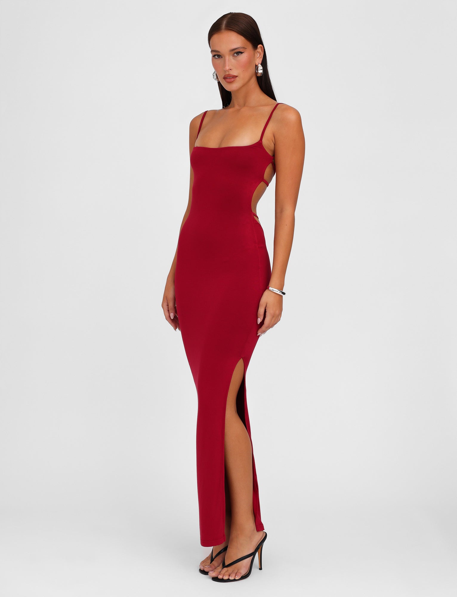 A woman stands against a white background wearing the TIGERMIST MINERVA MAXI DRESS - RED : BERRY RED, a sleeveless, full-length dress featuring side cutouts and a thigh-high slit. The bodycon silhouette highlights her figure while adjustable shoulder straps ensure the perfect fit. She accessorizes with dangling earrings, a bracelet, and black high-heeled sandals.