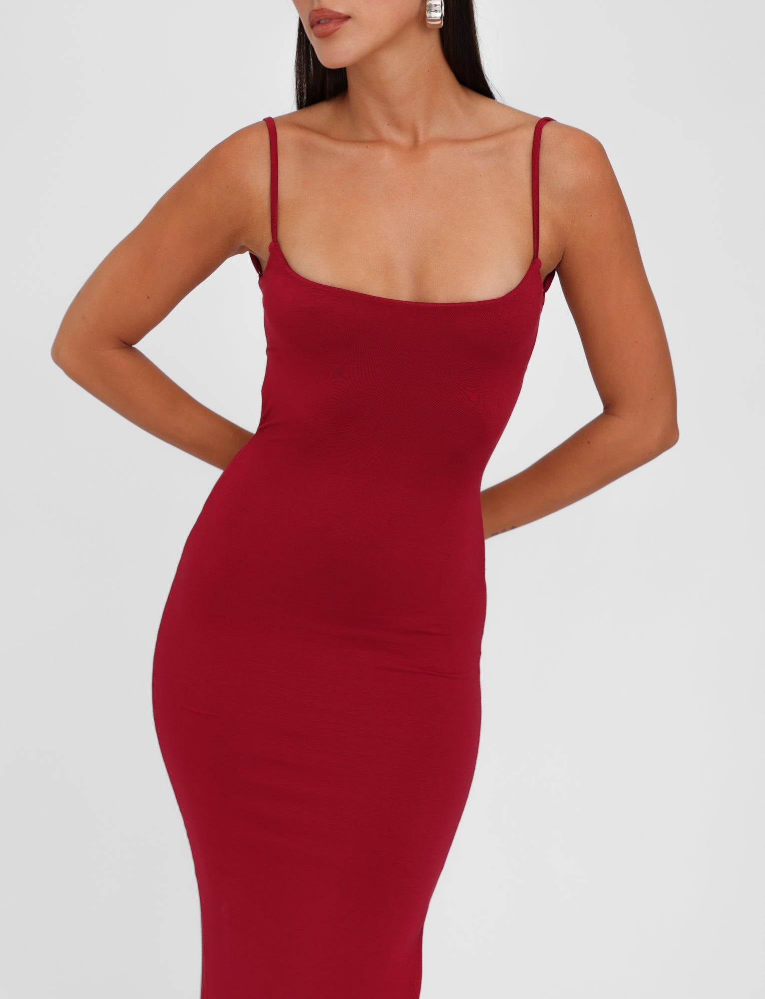 A woman wearing a Minerva Maxi Dress in Berry Red by Tigermist stands with her hands behind her back. The dress is form-fitting and sleeveless, featuring thin adjustable shoulder straps. She has long hair and is adorned with large square-shaped earrings against a plain white background.
