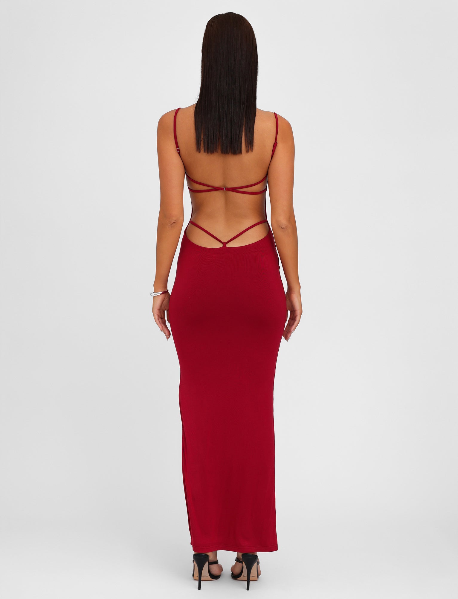 A woman faces away from the camera while wearing the TIGERMIST MINERVA MAXI DRESS in Berry Red—an elegant, long dress featuring a bodycon silhouette and adjustable shoulder straps. She has straight hair and complements her look with black high-heeled sandals against a plain white background.