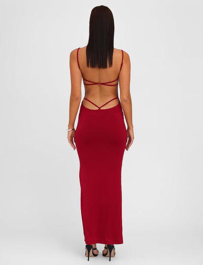 A woman faces away from the camera while wearing the TIGERMIST MINERVA MAXI DRESS in Berry Red—an elegant, long dress featuring a bodycon silhouette and adjustable shoulder straps. She has straight hair and complements her look with black high-heeled sandals against a plain white background.