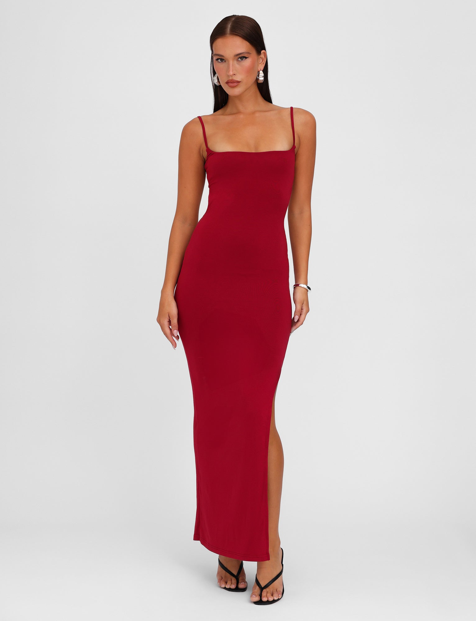 A woman stands against a plain white background wearing the MINERVA MAXI DRESS - RED : BERRY RED by TIGERMIST, a form-fitting, ankle-length red dress with adjustable shoulder straps. She has long, straight hair and is accessorized with earrings, a bracelet, and black heeled sandals.