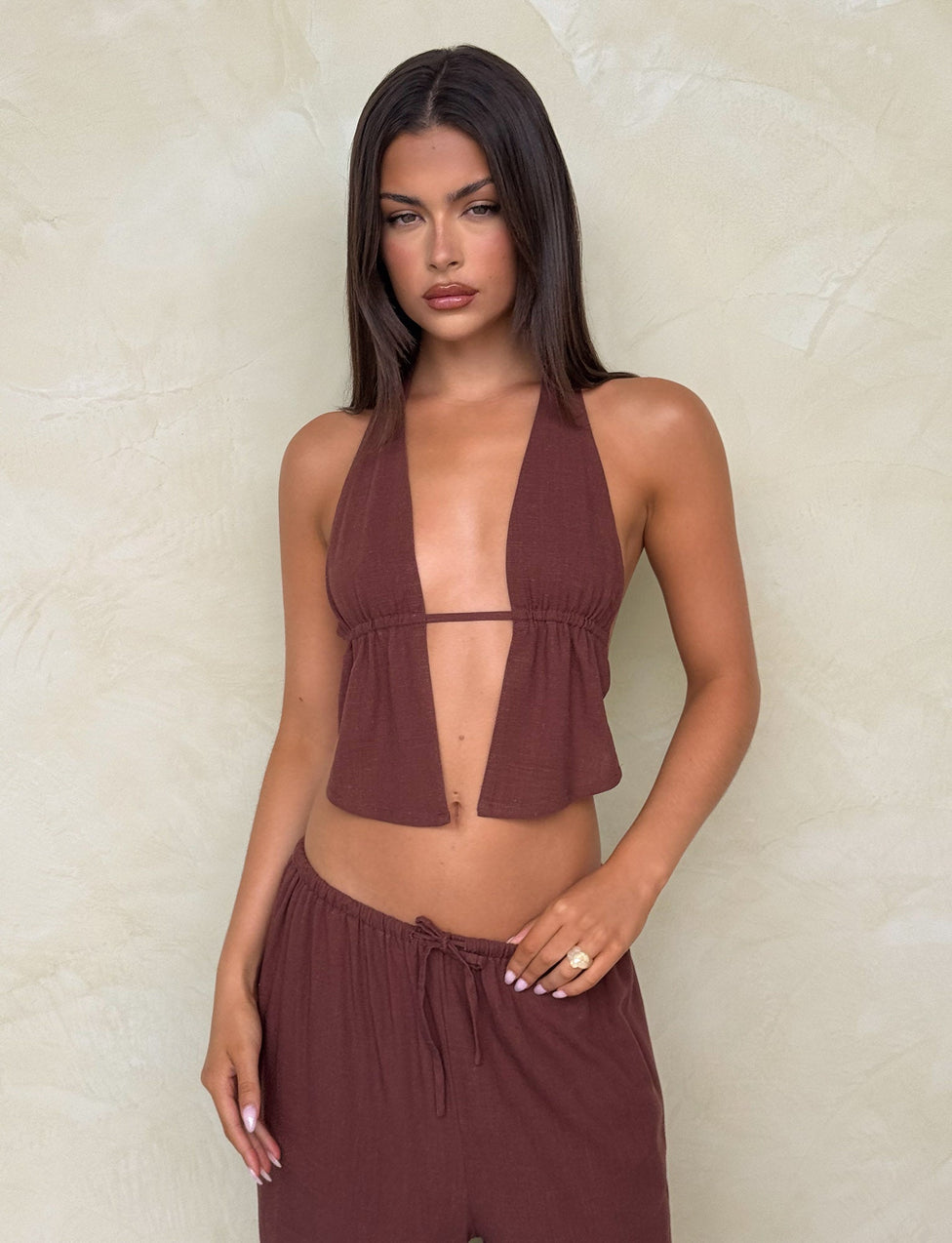 A woman with long dark hair wears the TIGERMIST LAINEY TOP - CHOCOLATE, a chocolate brown adjustable linen halter top, paired with matching pants. She stands against a light beige textured wall with a neutral expression and hand on her hip.