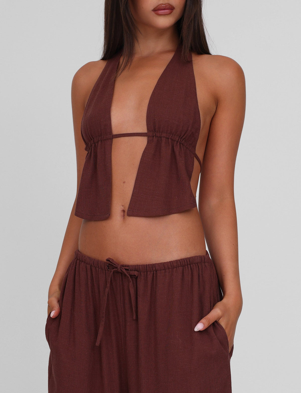 A woman wears the TIGERMIST LAINEY TOP - CHOCOLATE, a sleeveless, deep V-neck halter with open front and matching drawstring pants, hands in pockets. The background is plain and light-colored.
