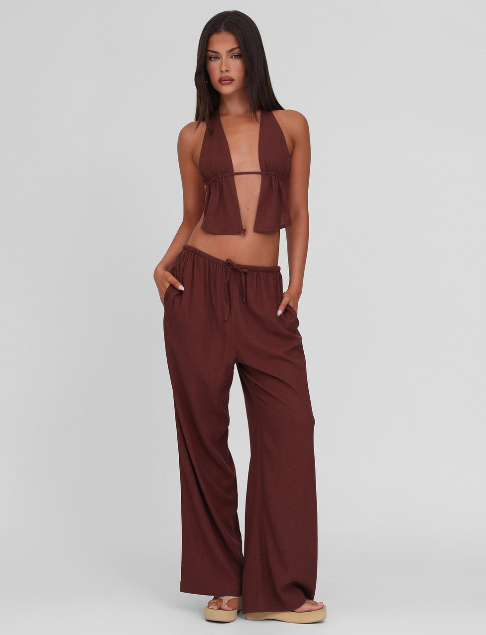 A woman with a neutral expression stands against a plain background wearing TIGERMIST’s LAINEY PANT in Chocolate—a wide leg linen style featuring functional side seam pockets and a mid-rise elastic waist—paired with a brown halter crop top and beige sandals.