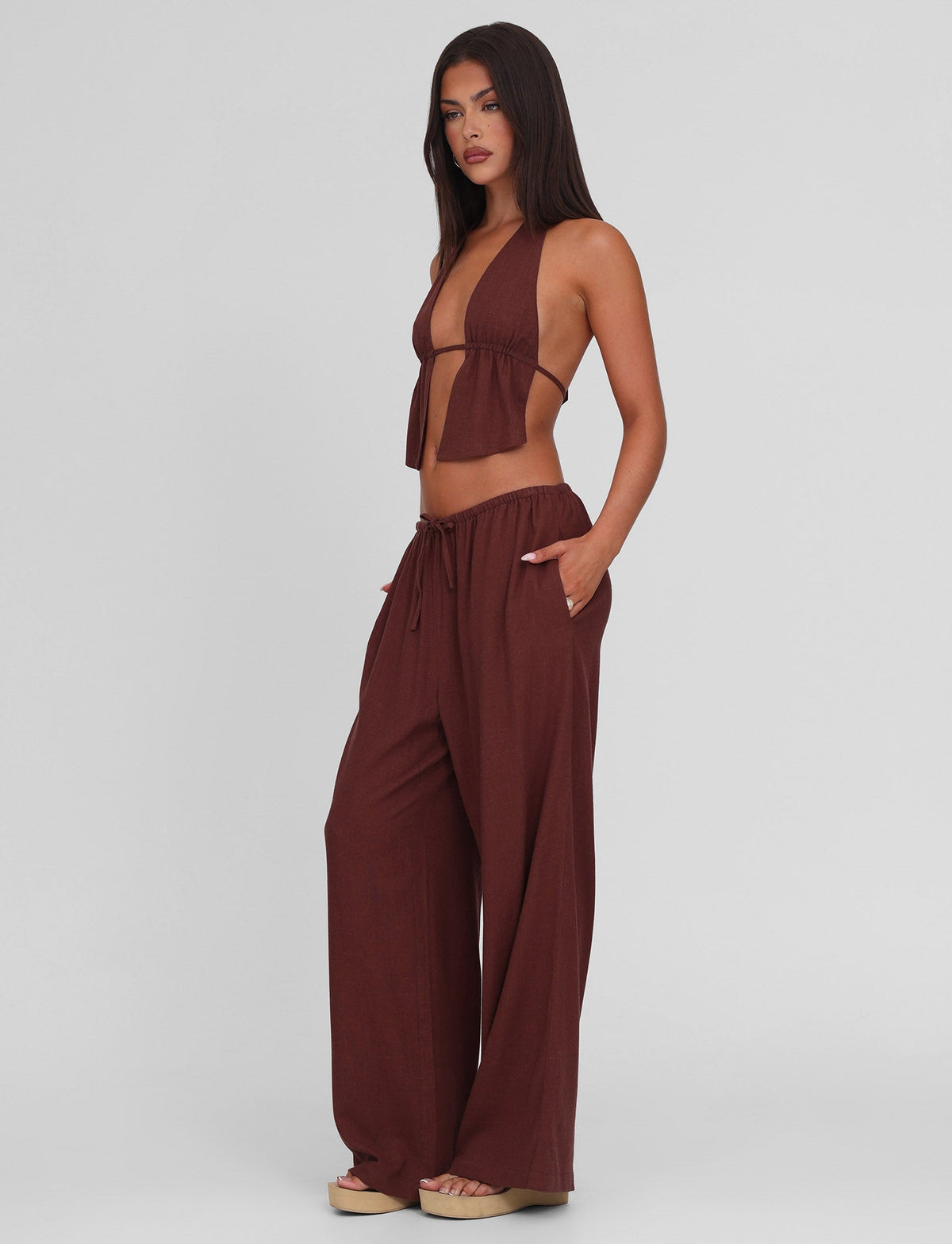 A woman wearing the TIGERMIST LAINEY PANT in Chocolate, featuring a mid-rise elastic waist, wide leg, and functional side seam pockets, stands against a plain light gray background.