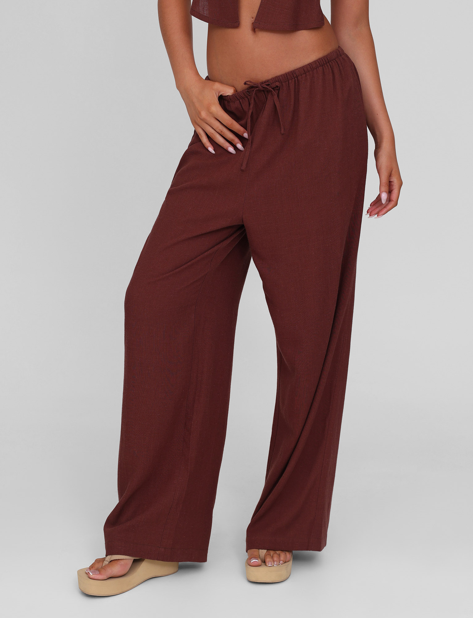 A person models the TIGERMIST LAINEY PANT in chocolate, featuring wide legs, a mid-rise elastic waist, and side seam pockets, paired with beige platform sandals, posing with one hand on their hip against a plain gray background.