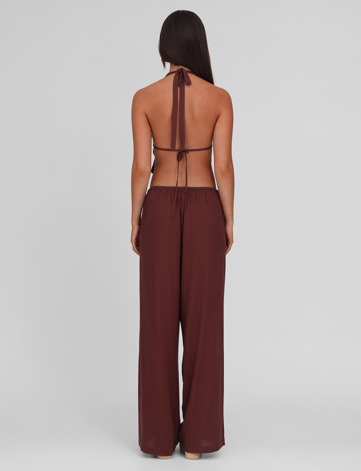 A woman with long dark hair stands facing away against a plain light gray background, wearing the TIGERMIST LAINEY PANT - CHOCOLATE, featuring wide legs and functional side seam pockets paired with a matching brown halter top.