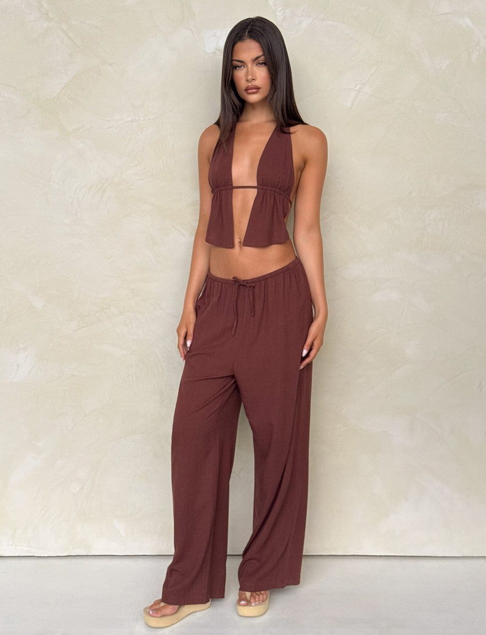 A woman stands against a textured beige wall, wearing the TIGERMIST LAINEY PANT in chocolate—a wide leg linen style with mid-rise elastic waist and side seam pockets—paired with a sleeveless brown halter top and beige platform sandals.