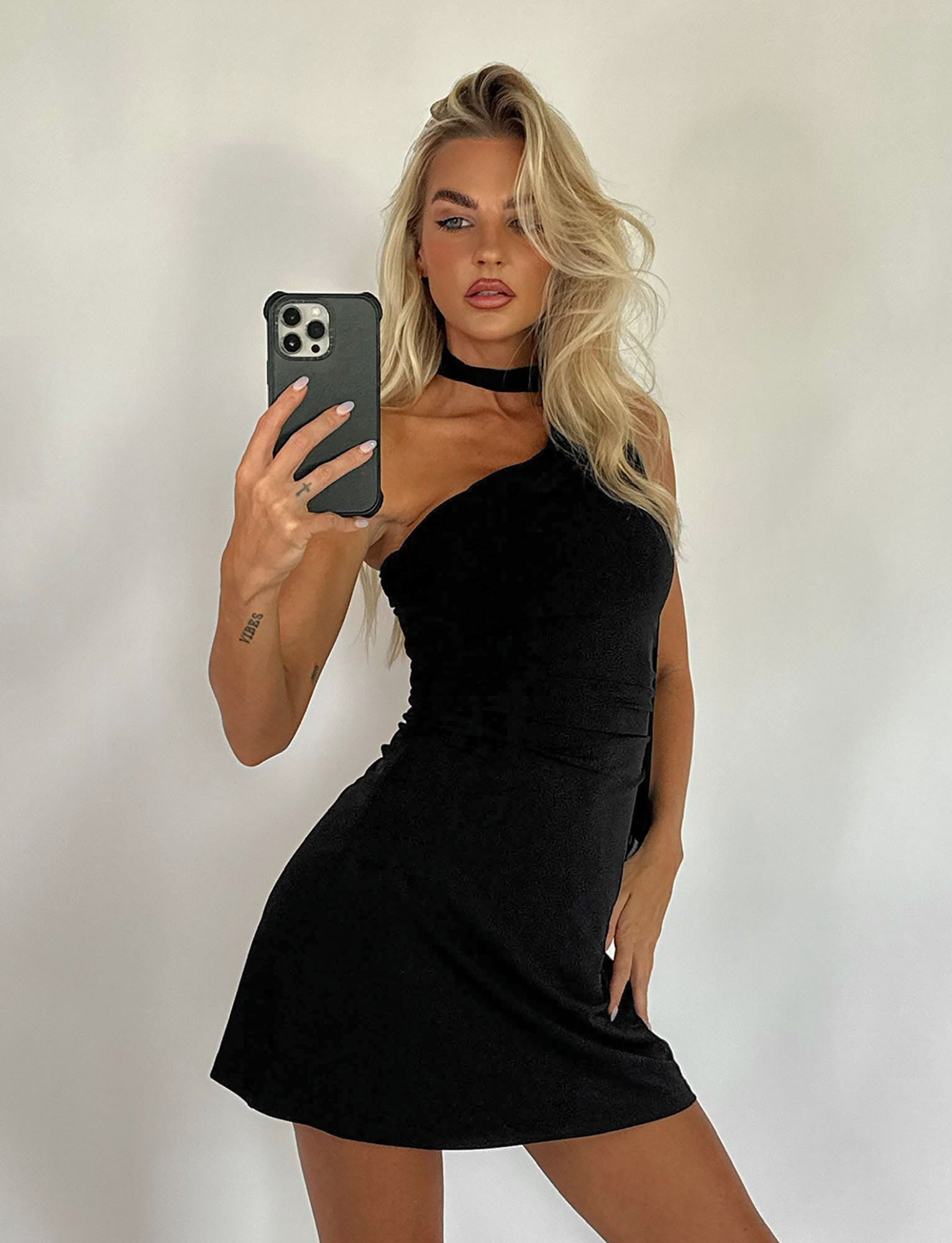 A woman with long blonde hair is taking a mirror selfie using her smartphone. She is wearing the sleek, black ALLIRA DRESS from TIGERMIST and stands against a plain, light-colored background.